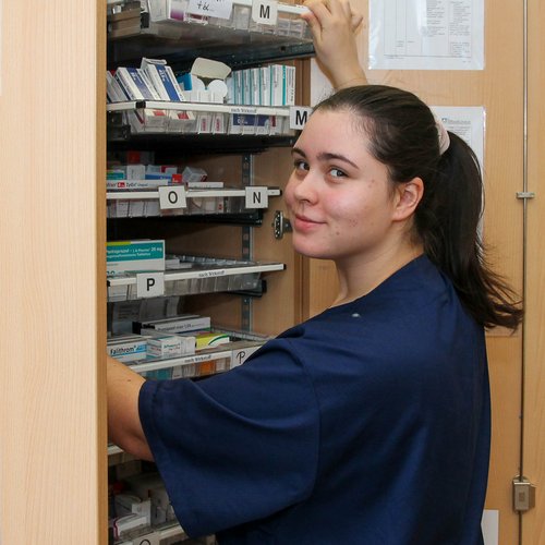 Eine Schülerin steht am geöffneten Medikamentenschrank und guckt in die Kamera.