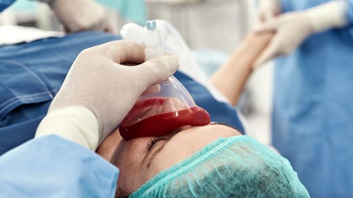 Bild von Frau auf einem OP-Tisch. Medizinisches Personal platziert eine Narkose-Maske über ihren Mund und ihre Nase.